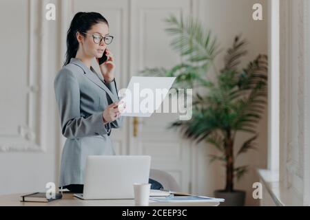 Serious brunette female employee stands near desktop, holds paper documents, talks via smartphone, wears formal clothes and spectacles, discusses busi Stock Photo