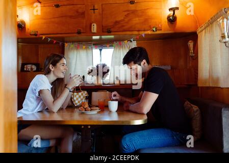 Side view of young man and Woman smiling and having good breakfast while sitting at table inside cozy caravan Stock Photo