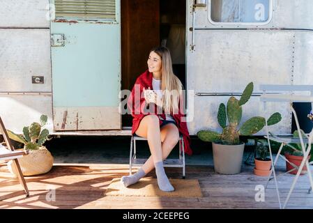 Pretty young lady with mug of hot beverage smiling and looking away while sitting on shabby camper in countryside Stock Photo