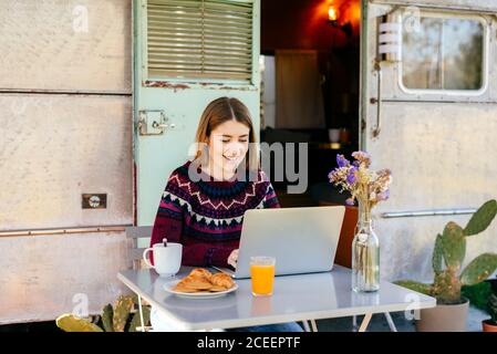 Cute young lady smiling and browsing modern laptop while sitting at table with breakfast food near shabby van Stock Photo