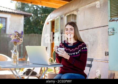 Cute young lady smiling and browsing modern laptop while sitting at table with breakfast food near shabby van Stock Photo