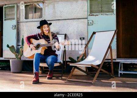 Beautiful young lady in stylish outfit sitting on chair near aged van and playing acoustic guitar Stock Photo