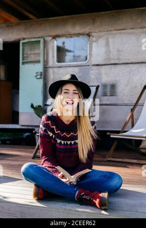 Charming young Woman in stylish outfit reading interesting book while sitting on terrace near aged camper Stock Photo