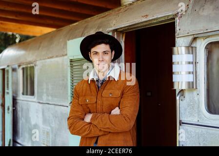 Handsome young man with crossed arms smiling and looking at camera while standing near entrance of aged caravan Stock Photo