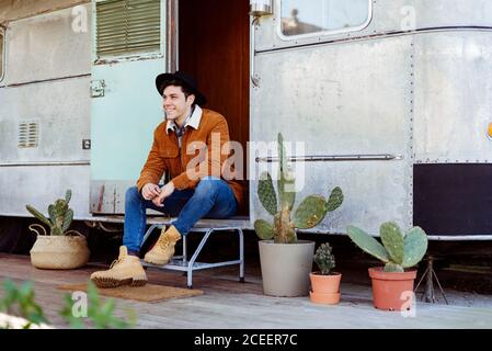 Attractive young man in stylish outfit smiling and looking away while sitting near entrance of shabby van Stock Photo