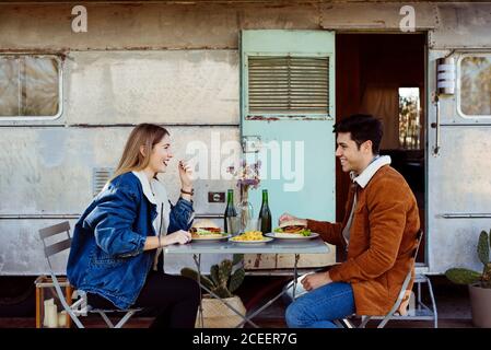 Side view of young man and Woman smiling and looking at each other while having delicious romantic dinner near shabby caravan Stock Photo