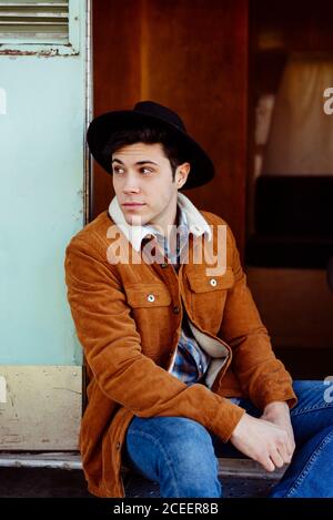 Attractive young man in stylish outfit smiling and looking away while sitting near entrance of shabby van Stock Photo