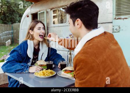 Side view of young man and Woman smiling and looking at each other while having delicious romantic dinner near shabby caravan Stock Photo