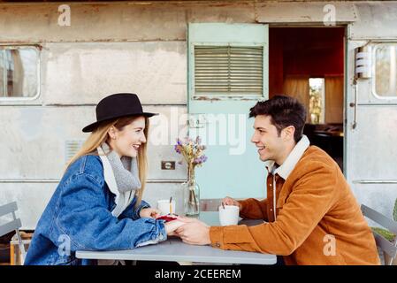 Young man and Woman smiling and looking at each other while sitting at table with hot beverage and piece of birthday cake near weathered camper Stock Photo