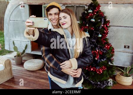 Young couple smiling and posing for selfie while standing near amazing Christmas tree outside retro camper Stock Photo