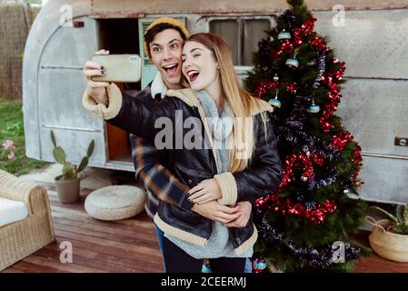 Young couple smiling and posing for selfie while standing near amazing Christmas tree outside retro camper Stock Photo