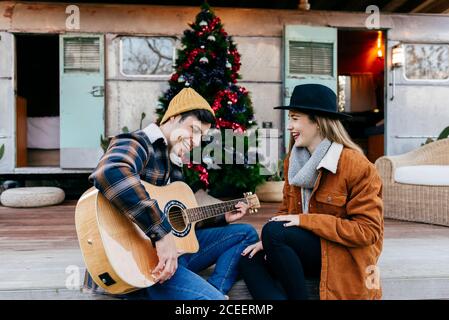 Handsome young man smiling and playing guitar for lovely girlfriend while sitting near Christmas tree and weathered trailer Stock Photo
