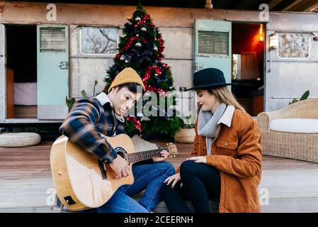 Handsome young man smiling and playing guitar for lovely girlfriend while sitting near Christmas tree and weathered trailer Stock Photo