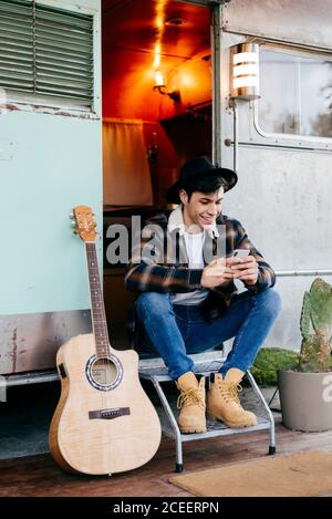 Handsome young man in stylish outfit smiling and using modern smartphone†while sitting in old caravan doorway near acoustic guitar Stock Photo