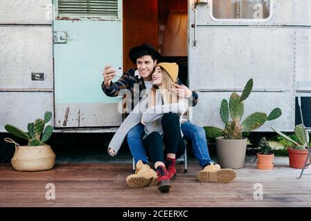 Handsome young man and beautiful Woman smiling and posing for selfie while sitting near weathered caravan Stock Photo