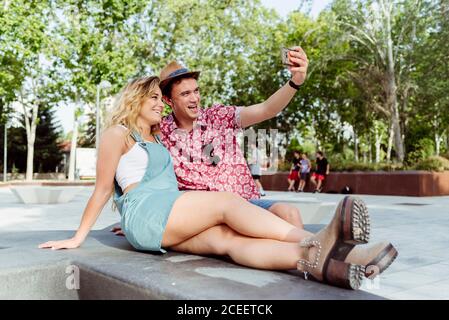 Cheerful young man and Woman sitting on modern bench and taking selfie Stock Photo