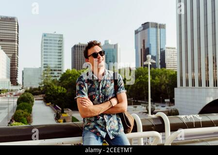 Cheerful young man in sunglasses leaning on handrail and looking away in the city Stock Photo