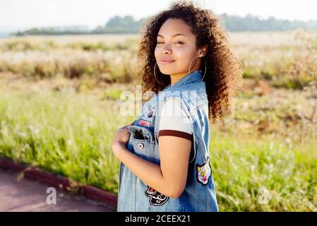 Charming African-American female teenager looking at camera while walking in amazing countryside on sunny day Stock Photo