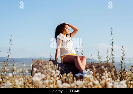 Lovely ethnic Woman sitting on big log and enjoying weather while spending time in nature Stock Photo