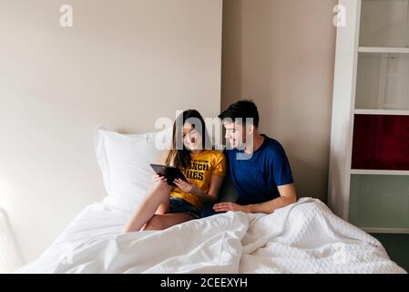 Cheerful young man and Woman lying in bed together and using digital tablet at home. Stock Photo