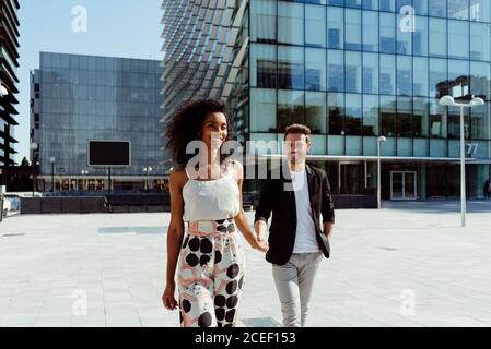 Pretty black Woman and Caucasian man smiling and holding hands while walking on city street together on sunny day Stock Photo