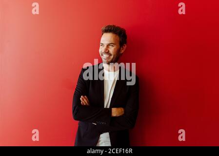 Handsome young man in elegant outfit smiling and looking at camera while standing on bright red background Stock Photo