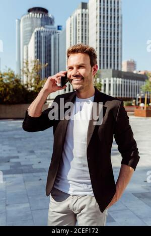 Attractive young man smiling using smartphone while standing on street of modern city on sunny day Stock Photo