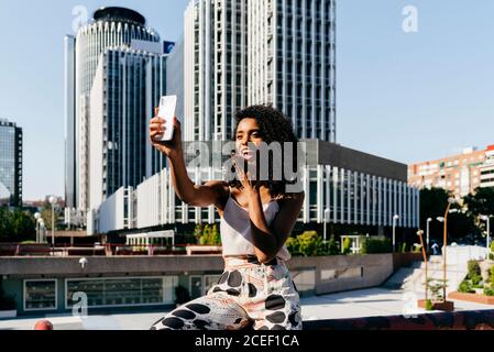 Beautiful African-American Woman laughing and posing for selfie while sitting on border on city street on sunny day Stock Photo
