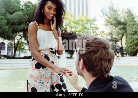Caucasian man putting engagement ring on finger of black Woman while proposing to her near fountain in park Stock Photo