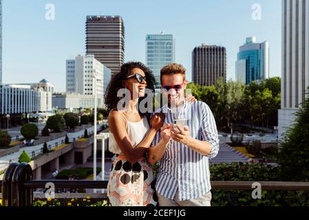 Beautiful black Woman and handsome Caucasian man using smartphone while standing on background of modern city Stock Photo