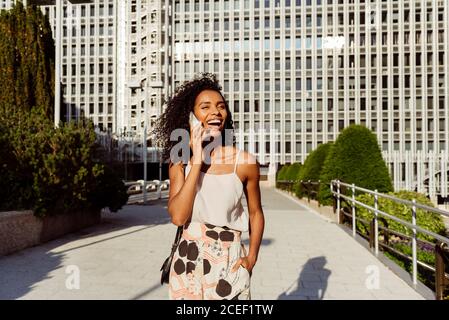 Attractive African-American lady laughing and talking on smartphone while walking on city street on sunny day Stock Photo