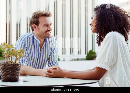 Side view of pretty African-American Woman and handsome Caucasian man holding hands and looking at each other while sitting at table in outdoor cafe Stock Photo