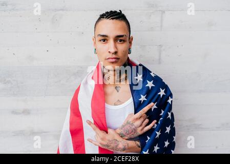 Hispanic tattooed man with piercing wrapping himself up in flag of united states and looking at camera on grey textured background Stock Photo