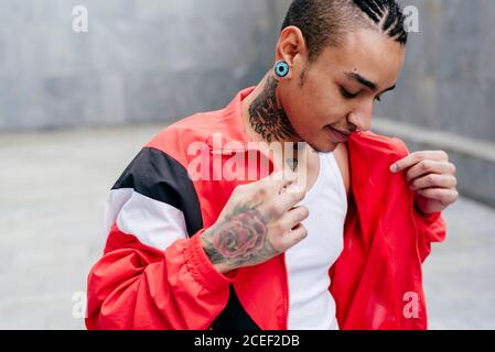 Smiling hipster with piercing and tunnel in ears holding red casual jacket in hands on grey blur background Stock Photo