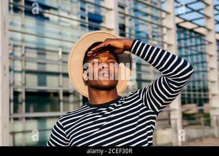 Attractive African-American man in hat keeping hand near forehead and looking at distance while standing on street Stock Photo