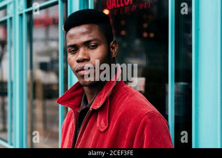Stylish african male putting hands in pocket of red jumper standing at entrance to cafe Stock Photo