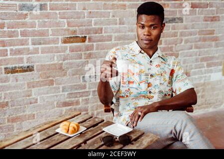 Handsome African-American man in stylish outfit drinking hot beverage and looking at camera while sitting at small table Stock Photo