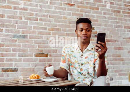 Handsome black man drinking hot beverage and browsing smartphone while sitting at small table near brick wall Stock Photo