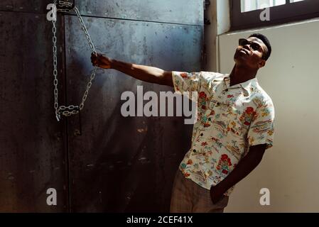 Relaxed black man in stylish outfit grasping chain and leaning back while standing in stylish loft Stock Photo