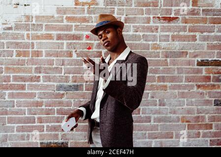 Handsome African-American man in elegant outfit holding deck of playing card and throwing up two dices while standing near brick wall Stock Photo