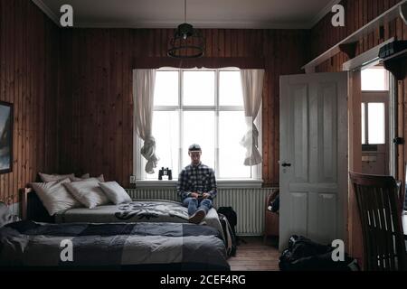 Adult man sitting and resting on bed while using smartphone at home in Iceland Stock Photo