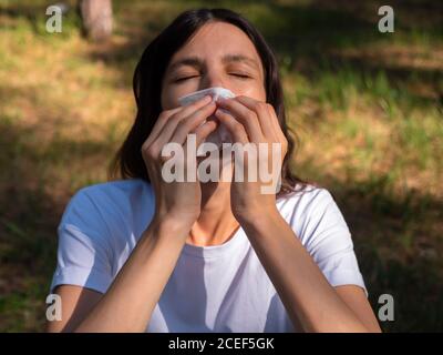 Woman with eyes closed suffering from grass pollen allegry sneezes in paper tissue on a sunny day in park. Allergic running nose concept. Stock Photo