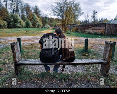 Back view of father with backpack and daughter with long loose hair sitting in the country on a bench with their heads on each other's shoulders. Stock Photo