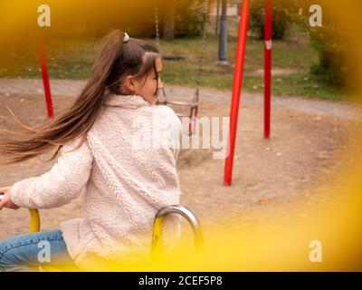 View through railings over happy smiling teenage girl in a faux fur coat and with long ponytail spinning at a merry-go-round at children playground. Stock Photo