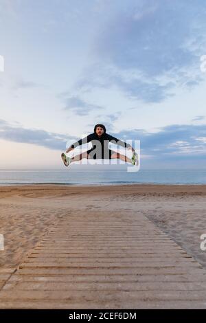 Handsome man in sportswear jumping high during outdoor training on sandy beach Stock Photo