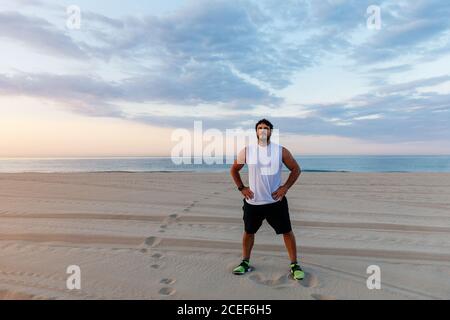 Positive handsome man in sportswear keeping hands on waist while standing on sandy beach during sunset Stock Photo