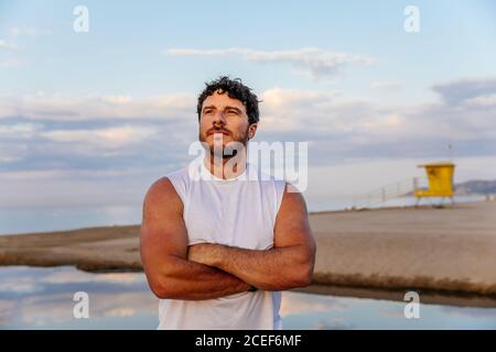 Bearded adult man in sportswear holding hand near forehead and looking at distance while standing on beach during outdoor training Stock Photo