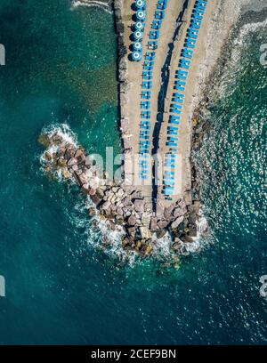 Aerial scenic view of rocky and sandy small peninsula with bright blue parasols and beach chairs surrounded by beautiful ocean Stock Photo