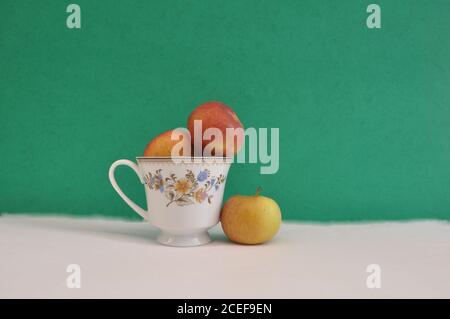 Coffee cup with floral motifs, with fresh apples inside and outside, conceptual photo of separation, segregation, on white base and green background Stock Photo