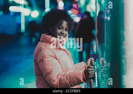 Side view of beautiful African American female smiling and looking at camera while playing arcade game in amusement park at night Stock Photo